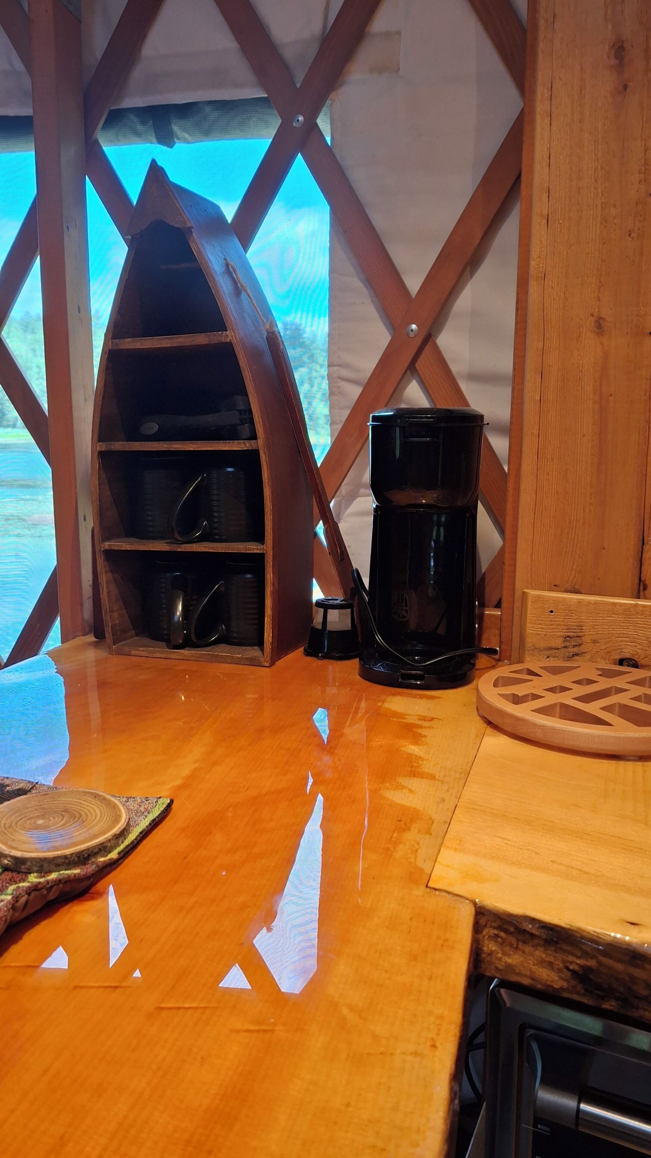Interior de madeira com uma cafeteira e canecas sobre uma mesa, uma prateleira de madeira com canecas, e uma vista pela janela.