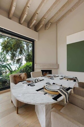 A round dining table with chairs, a fireplace, and a large window with a view of greenery and the sea.