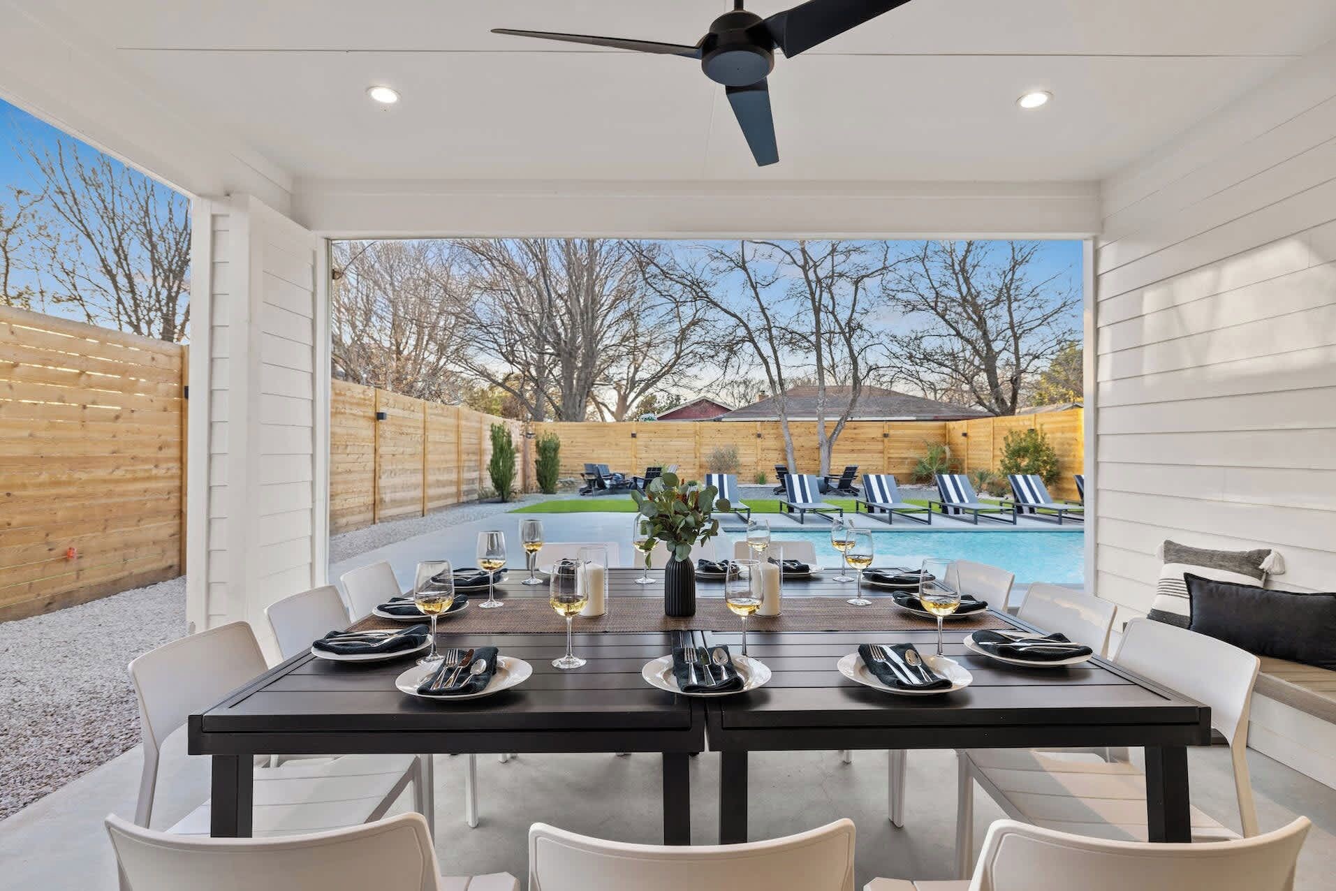 An outdoor dining area with a pool view, tables set for four, and a ceiling fan.