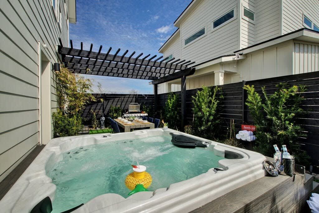 A hot tub in a backyard with a pergola overhead, surrounded by a fence and greenery.