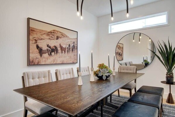A dining area with a long wooden table, cushioned chairs, and a large painting of horses. There is a round mirror on the wall and a potted plant on the side.