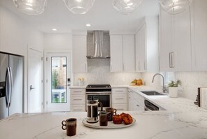 Eine moderne Küche mit weißen Schränken, einer Edelstahl-Dunstabzugshaube, einem eingebauten Ofen und einer großen Kochinsel mit Kaffeemaschine und Gebäck.