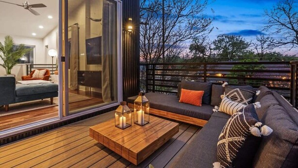 Un espace de vie extérieur moderne, doté d’une terrasse en bois, d’un canapé avec des coussins et d’une porte en verre donnant sur un espace intérieur.