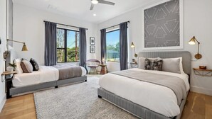 Une chambre avec deux lits, un ventilateur de plafond, de grandes fenêtres avec des rideaux, une œuvre d’art abstraite encadrée et des lampes de chevet.
