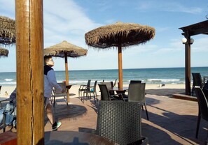 Un ristorante sulla spiaggia con posti a sedere all'aperto, tra cui tavoli e sedie, e ombrelloni di paglia che offrono ombra.