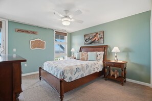 Une chambre à coucher comprenant un lit, une table de chevet, une commode, un ventilateur de plafond, une fenêtre et des éléments décoratifs sur les murs.