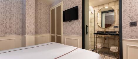 Una habitación de hotel con una cama grande, un televisor montado en la pared, un baño con ducha y un espejo, y un papel pintado estampado.