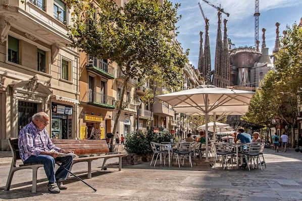 Un área de café al aire libre con mesas y sillas, rodeada de edificios con tiendas y balcones.