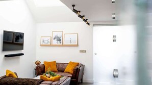 A modern living room with a high ceiling, a brown leather sofa with yellow cushions, a flat-screen TV, and framed artwork on the wall. - Stylish Cambridge 1-bed Home with free parking (Cambridge)