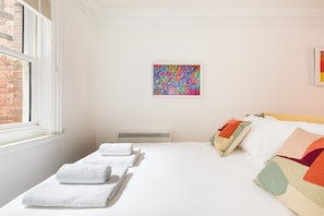 Quarto de hotel com cama bem arrumada, janela com vista para um edifício de tijolos e um quadro abstrato colorido na parede.