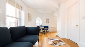 A modern living room with a dark sofa, a coffee table with a magazine, a dining area with chairs, and a large window. - Calm Leicester Sq 1BR (London)