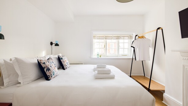 A neatly made bed with white linens and decorative pillows, a bedside table with a lamp, and a window with a view of the outside. - Calm Leicester Sq 1BR (London)