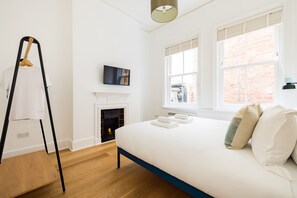 A bedroom with a bed, a wooden floor, a fireplace, and a window with a view of a brick building.