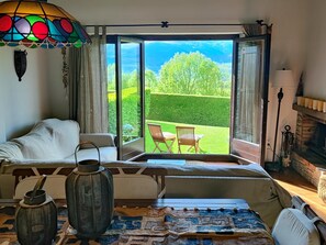 Une chambre avec un lit, une table sur laquelle se trouve une lanterne, une vue sur un jardin par une porte vitrée et une cheminée.