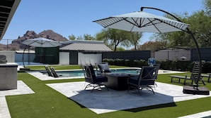 Un cortile con erba sintetica, piscina, mobili da esterno e un ombrellone grande.