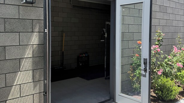 Un edificio moderno con facciata in mattoni grigi, ingresso con porta in vetro e una piccola area esterna con pavimento piastrellato e alcune piante.