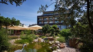 A courtyard with a pond, outdoor seating, and a multi-story building in the background. - Boutique Hotel Moselgarten (Bullay)