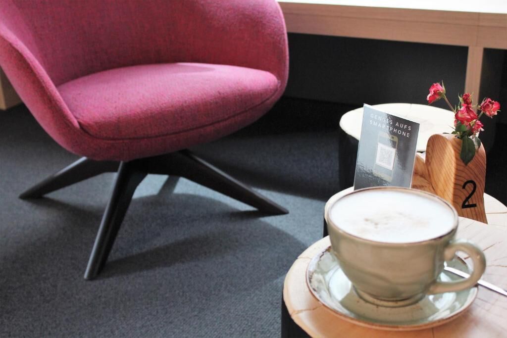 A modern room with a pink chair, a cup of coffee on a saucer, a book titled "Genius auf Smartphone", and a wooden table with a vase of flowers.
