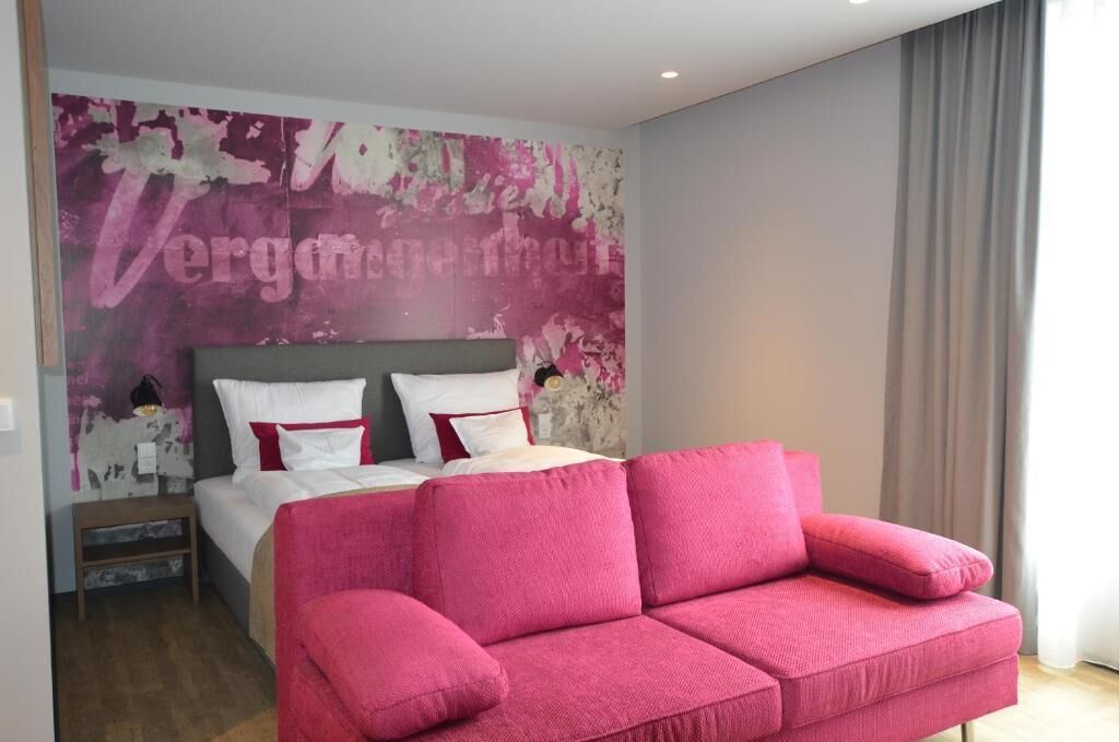 A bedroom with a bed, a pink sofa, and a wall with a large abstract painting.