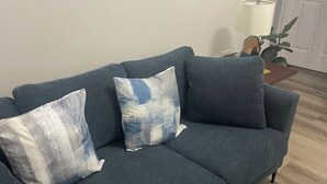 Un sofá azul oscuro con dos cojines a juego, una mesita de madera con una lámpara y una alfombra estampada azul.