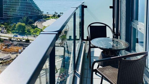 Een balkon met glazen hekwerk, twee rieten stoelen en uitzicht op zee en een gebouw in de verte.