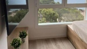 Un dormitorio con una ventana grande que ofrece vistas al mar y a árboles, una cama, un televisor montado y una planta pequeña en el suelo.