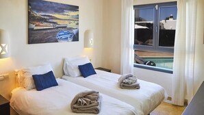 Hotellihuone, jossa on kaksi sänkyä, maalaus, jossa on kuvattuna veneitä, ikkuna, josta avautuu näkymä uima-altaalle, ja lamppu.