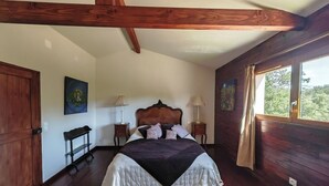 Een slaapkamer met een groot bed, houten balken, een raam met gordijnen en kunstwerken aan de wanden.