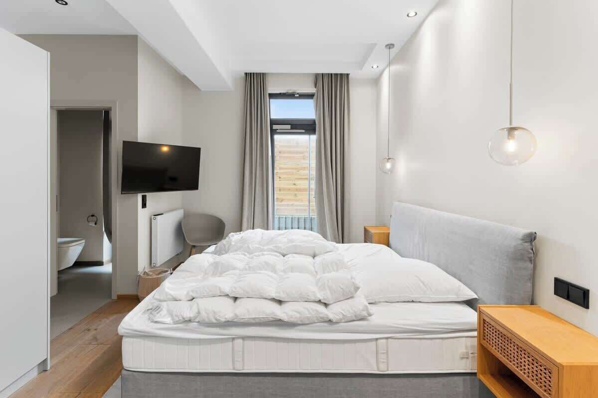 A modern bedroom with a bed, a TV mounted on the wall, a window with curtains, and a bedside table.