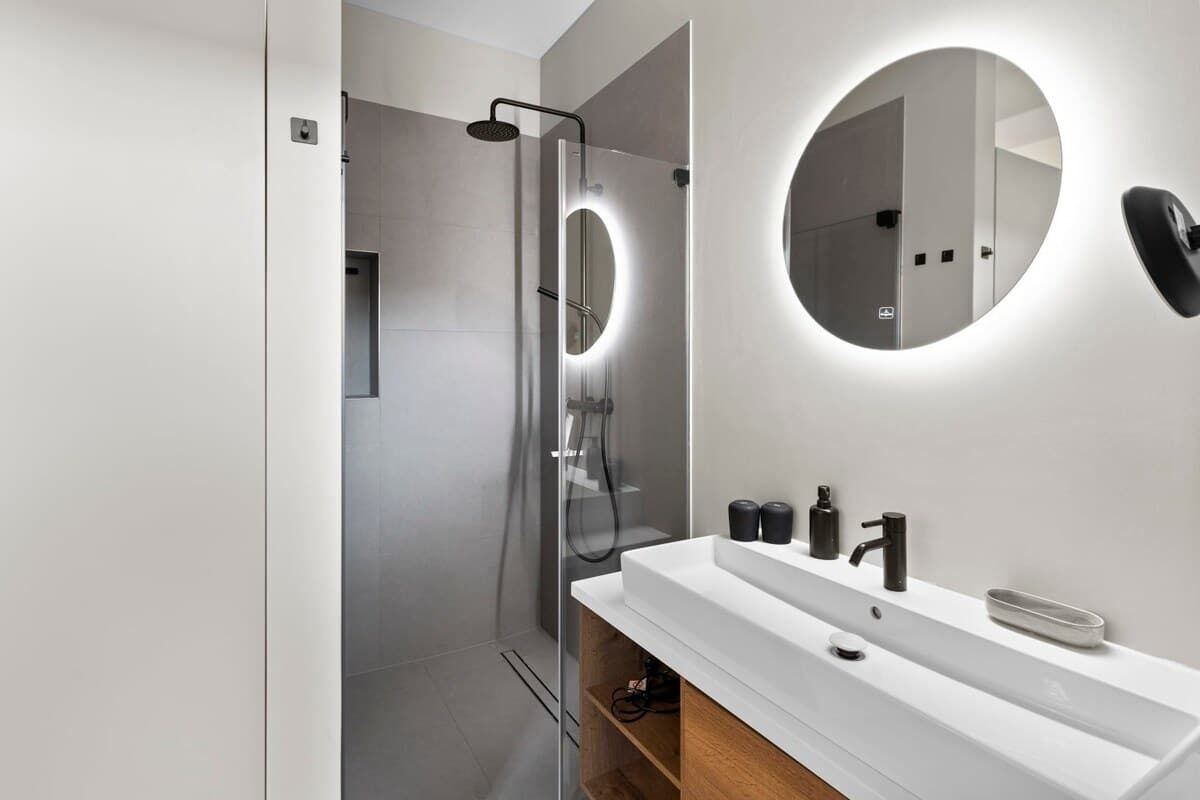 Une salle de bain moderne avec une douche à parois de verre, un lavabo blanc avec un miroir et un grand miroir rond fixé au mur.
