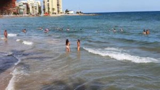 Uma praia com pessoas nadando e permanecendo na água, e edifícios ao fundo.