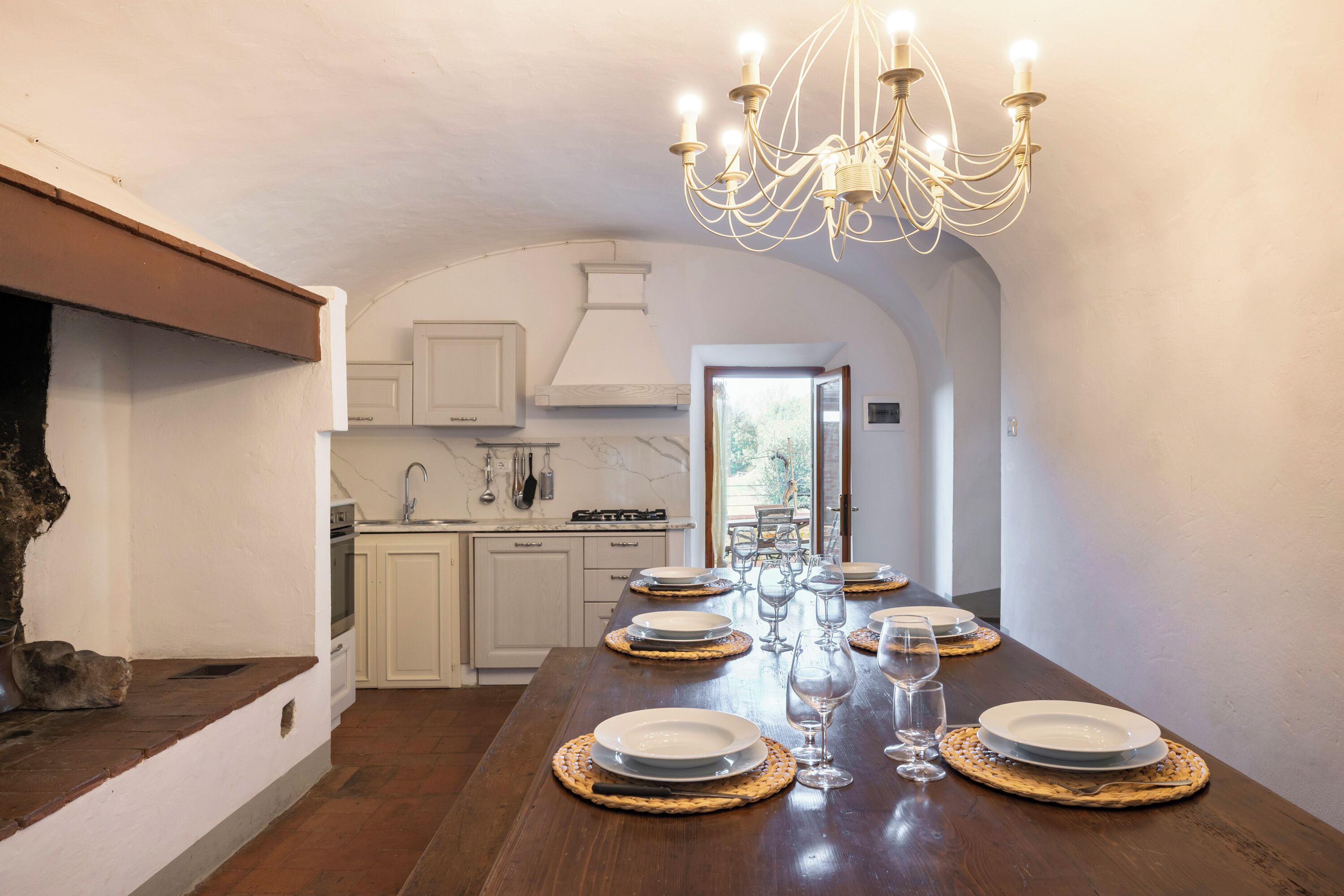 A long dining table set with plates and glasses, a chandelier, and a view into a kitchen area.
