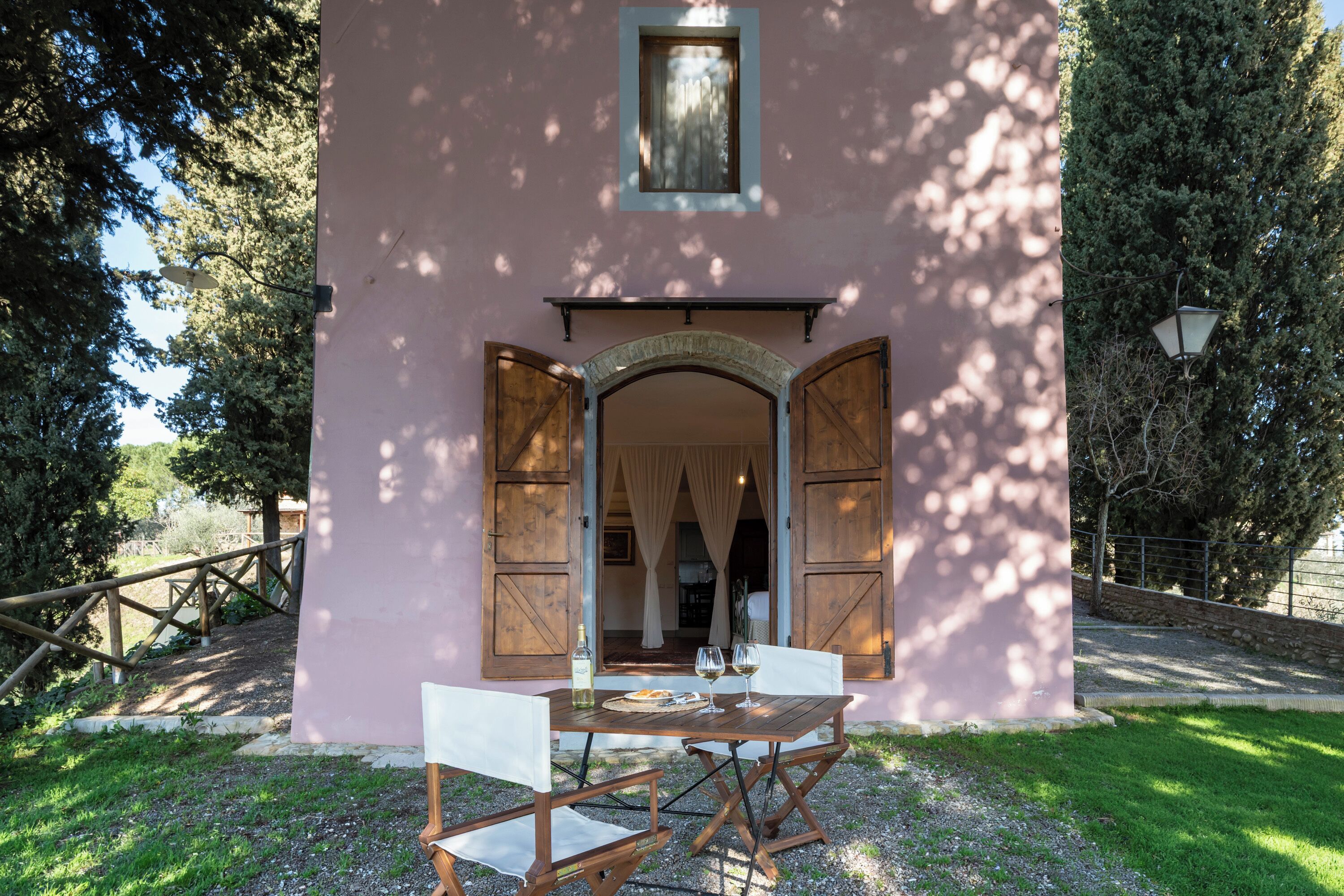 Um edifício rosa com uma arcada aberta, uma mesa com cadeiras e uma garrafa de vinho.