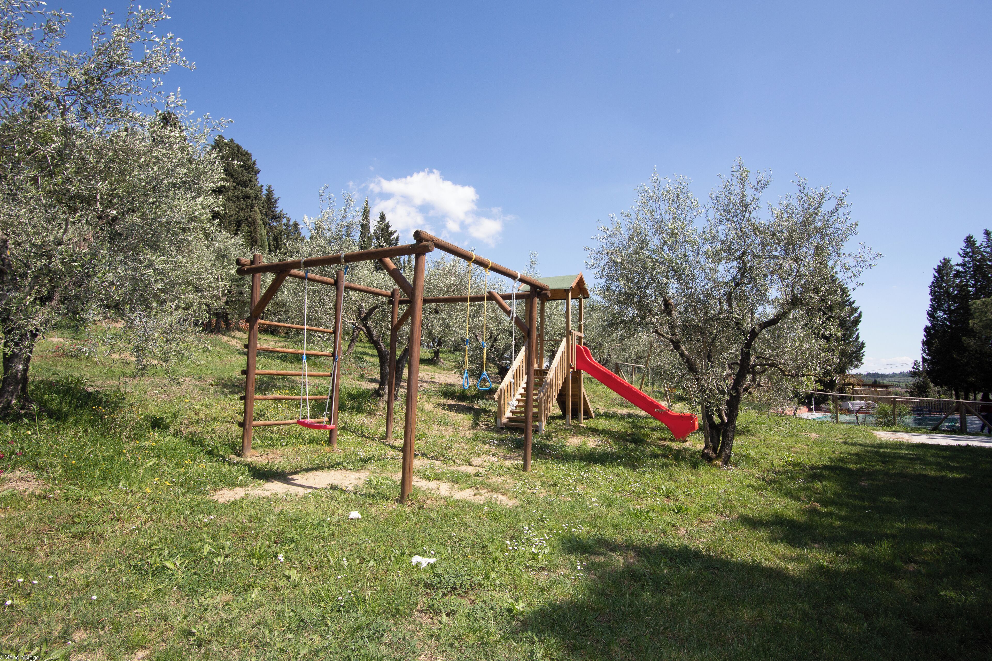 A playground with a wooden swing set and a red slide in a grassy area with trees.