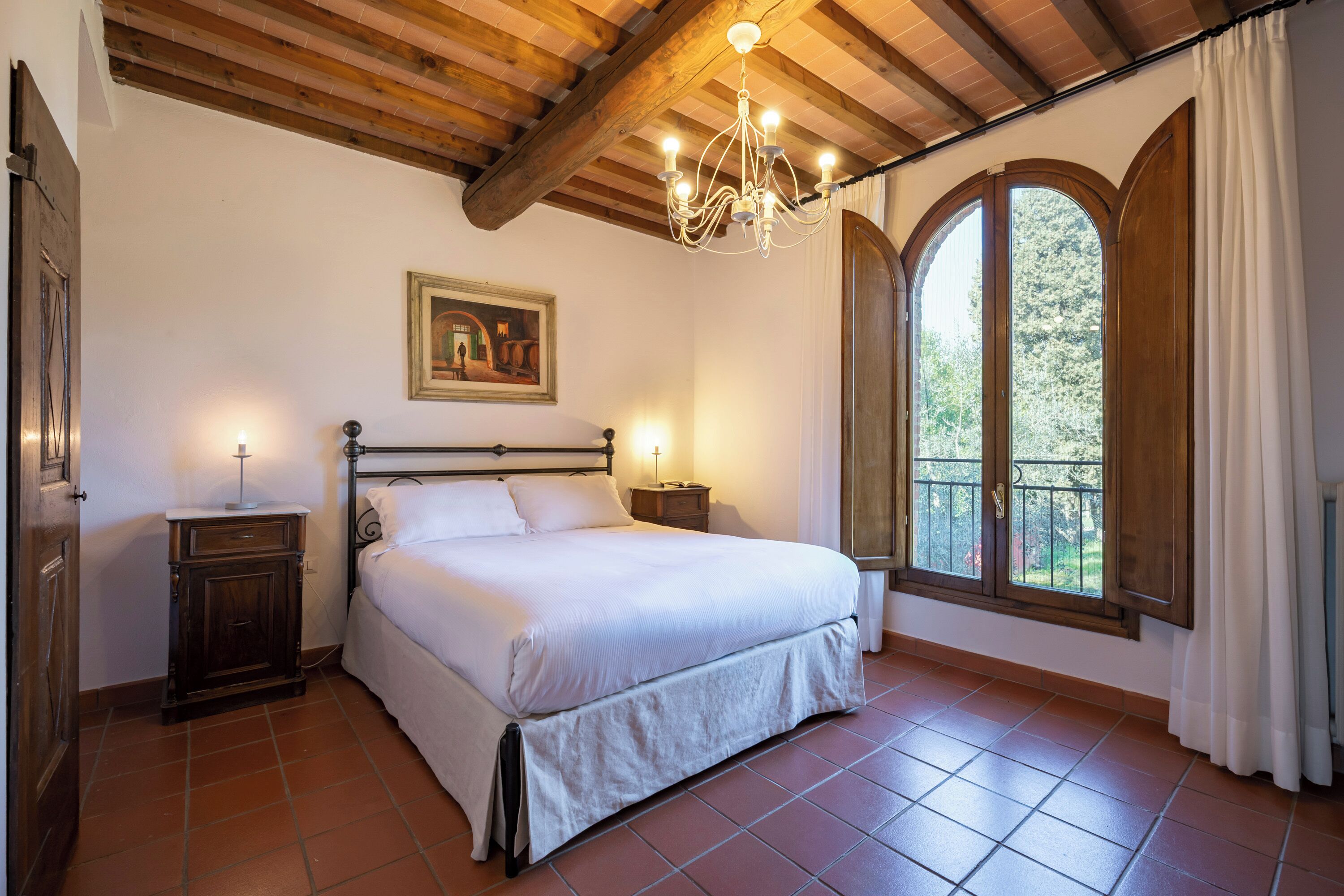 A bedroom with a bed, a nightstand, a chandelier, a painting, and a window with curtains.