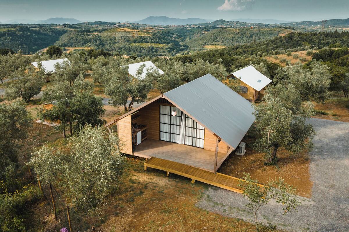 Eine moderne, A-förmige Hütte mit einem großen Schiebeglastür, umgeben von Olivenhainen und vor einer sanften Hügellandschaft gelegen.