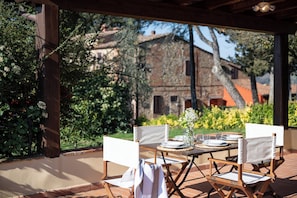 Zona pranzo all'aperto con tavolo imbandito per quattro, circondata da sedie in legno, e vista su un edificio rustico sullo sfondo.