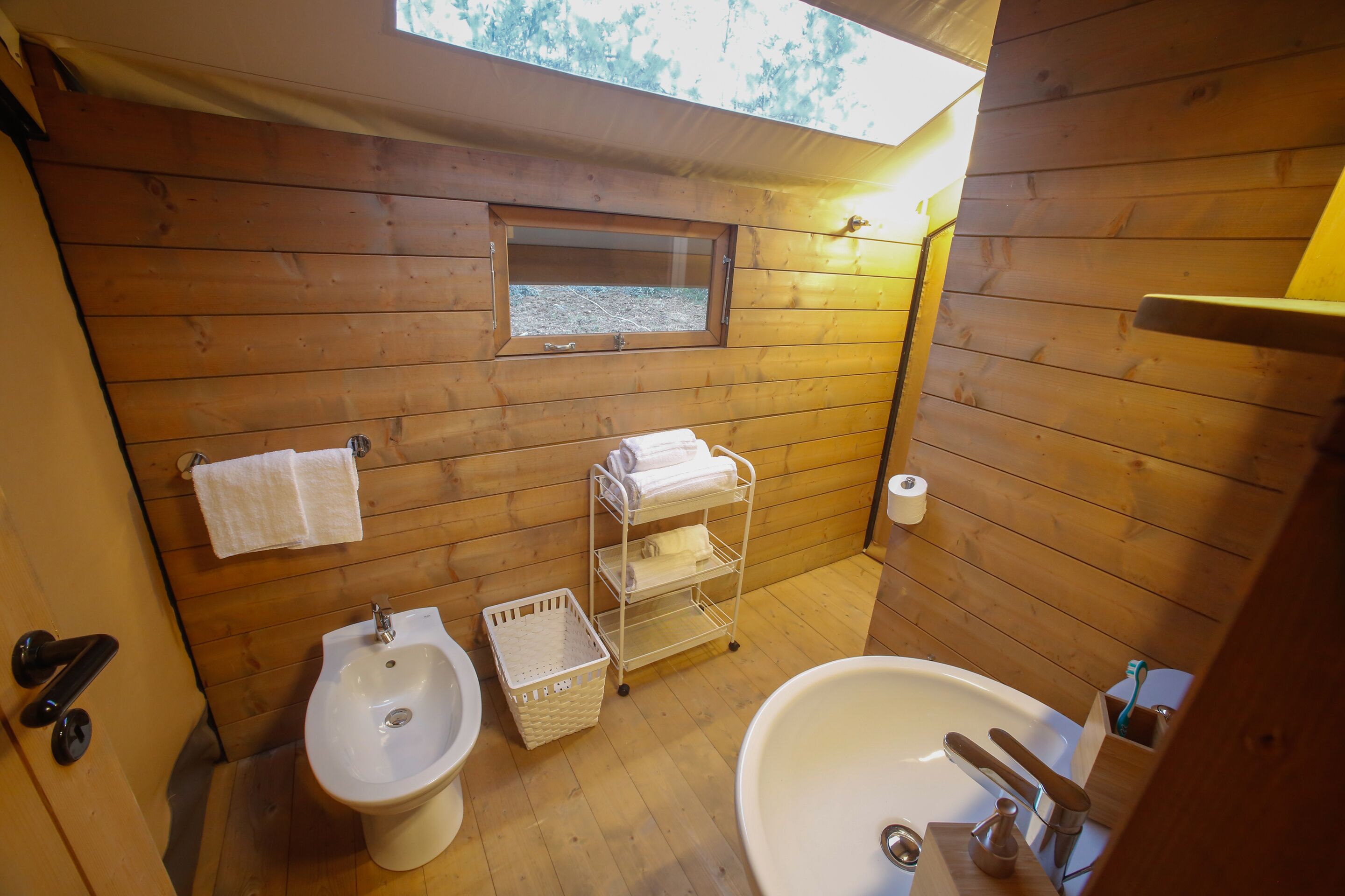 Een houten badkamer in een vakantiehuisje, met een hygiëneschuimer, wastafel en een plank met handdoeken.