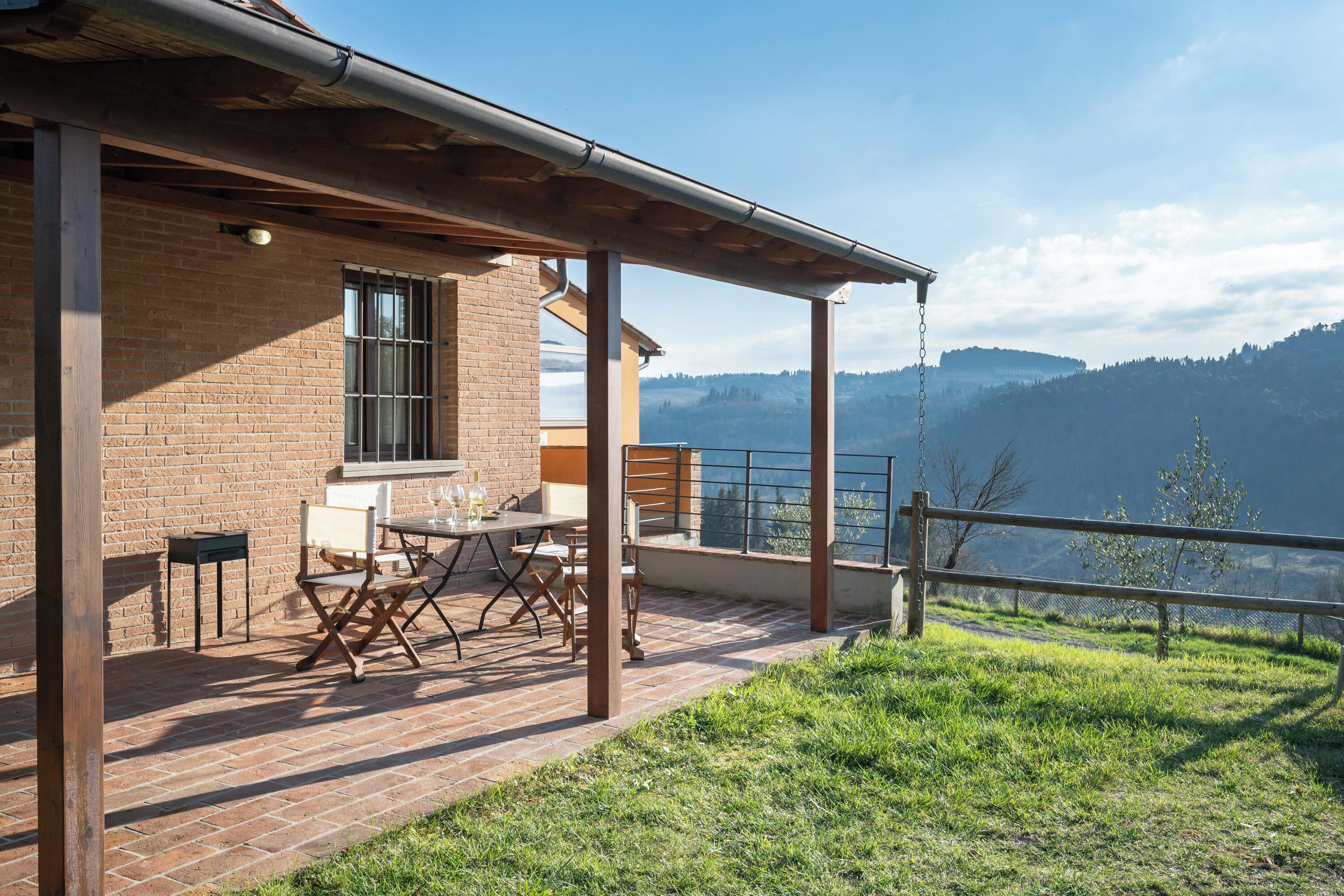 Ein überdachter Außenbereich mit Tisch und Stühlen, ein Backsteinhaus und Blick auf eine gebirgige Landschaft.