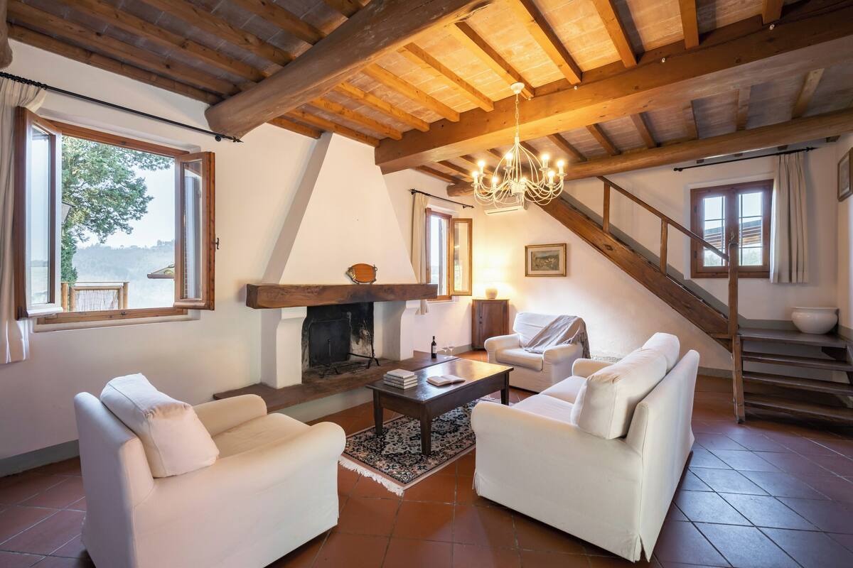 Ein geräumiges Wohnzimmer mit Kamin, weißen Sofas, einer Holzbalkendecke und einer Treppe.