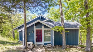 A small, single-story cabin with a red door and a blue exterior, surrounded by trees. - Margaritaville house next to pool (Osage Beach)