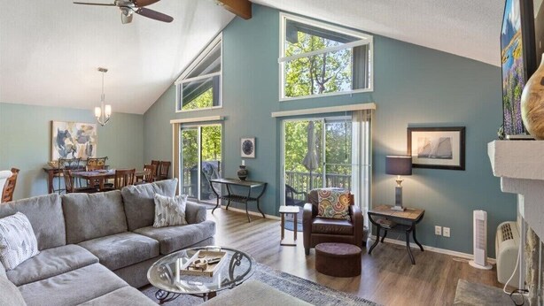 A spacious living room with a large gray sectional sofa, a glass coffee table, and a ceiling fan. There are multiple windows providing natural light and a view of the outdoors. - Margaritaville house next to pool (Osage Beach)