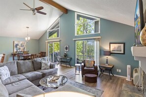 A spacious living room with a large gray sectional sofa, a glass coffee table, and a ceiling fan. There are multiple windows providing natural light and a view of the outdoors. - Margaritaville house next to pool (Osage Beach)