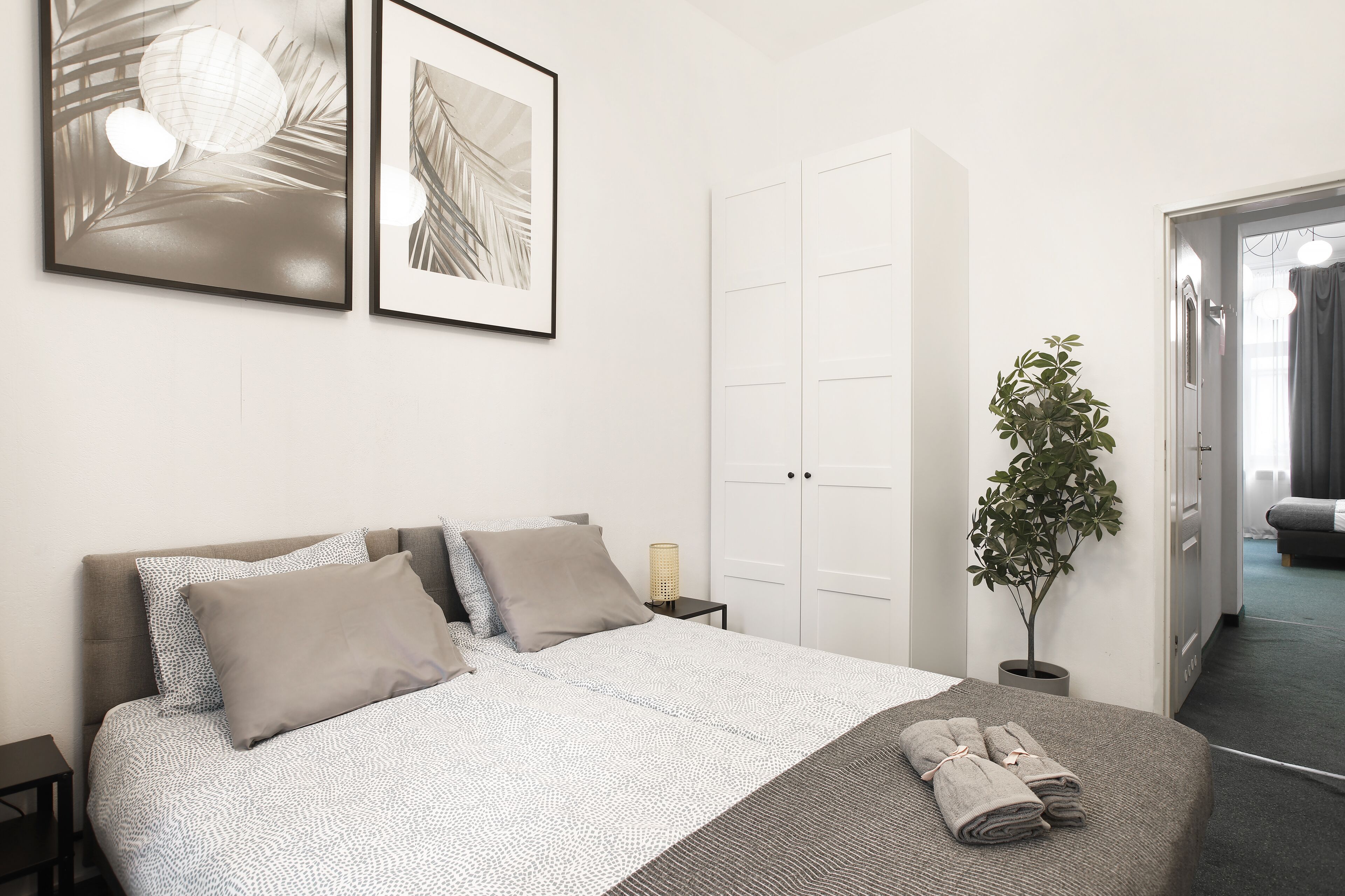 A neatly arranged bedroom with a bed, pillows, a bedside lamp, a potted plant, and framed artwork on the wall.
