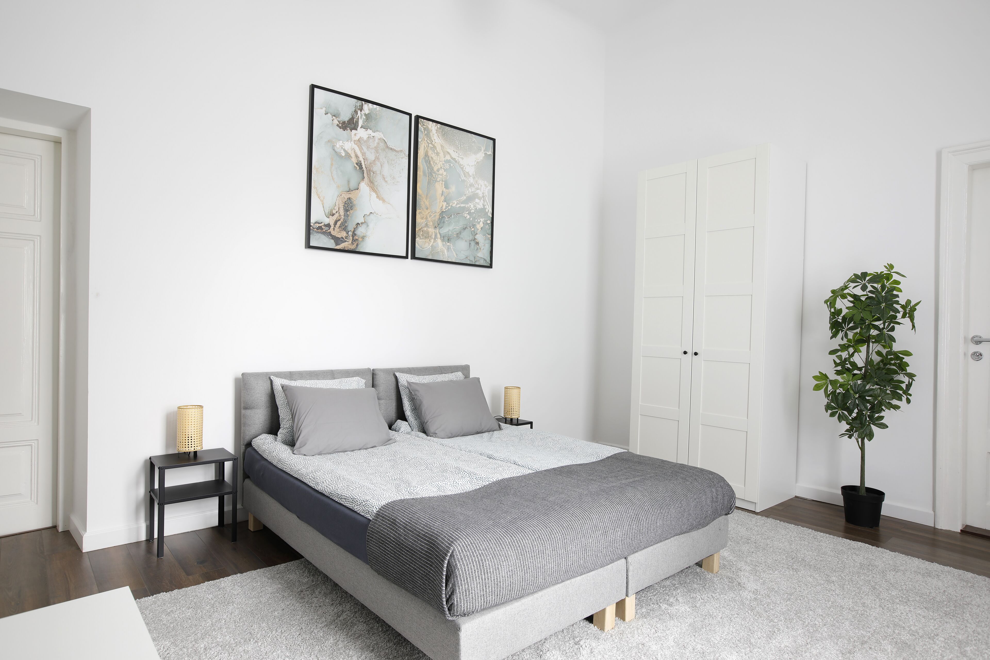 A modern bedroom with a grey bed, white walls, and a large potted plant.