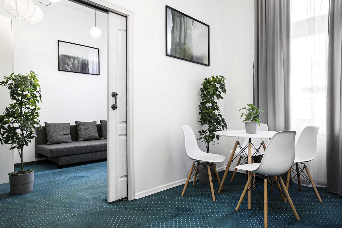 Ein modernes Zimmer mit einem grauen Sofa, weißen Stühlen und einem kleinen Tisch. Zwei Blumentöpfe und ein gerahmtes Bild sind an der Wand zu sehen.