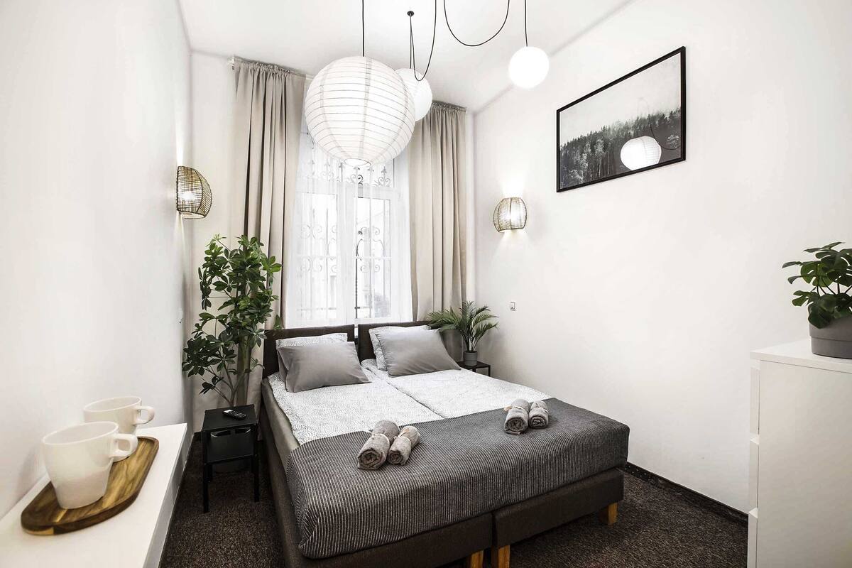 Ein modernes Schlafzimmer mit Bett, Nachttischen, einem Fenster mit Vorhängen, einem Wandbild und einem Topfpflanzen.