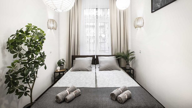 A minimalist hotel room with a large bed, two bedside tables, a potted plant, and a view of the city through sheer curtains. - Bedrouse Czapskich 5 - Old Town (Kraków)