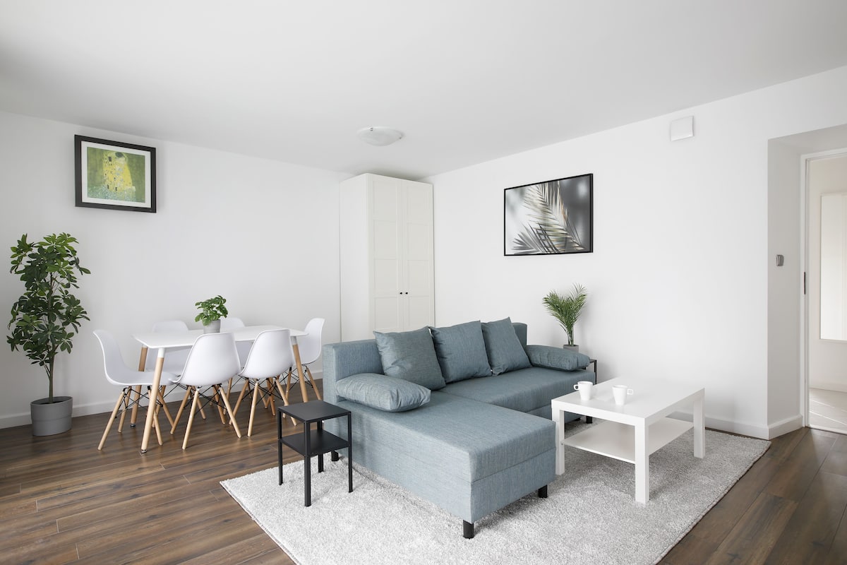 Ein modernes Wohnzimmer mit einem grauen Ecksofa, einem weißen Esstisch und Stühlen. Es gibt einen weißen Schrank und einen kleinen Tisch mit zwei Tassen.