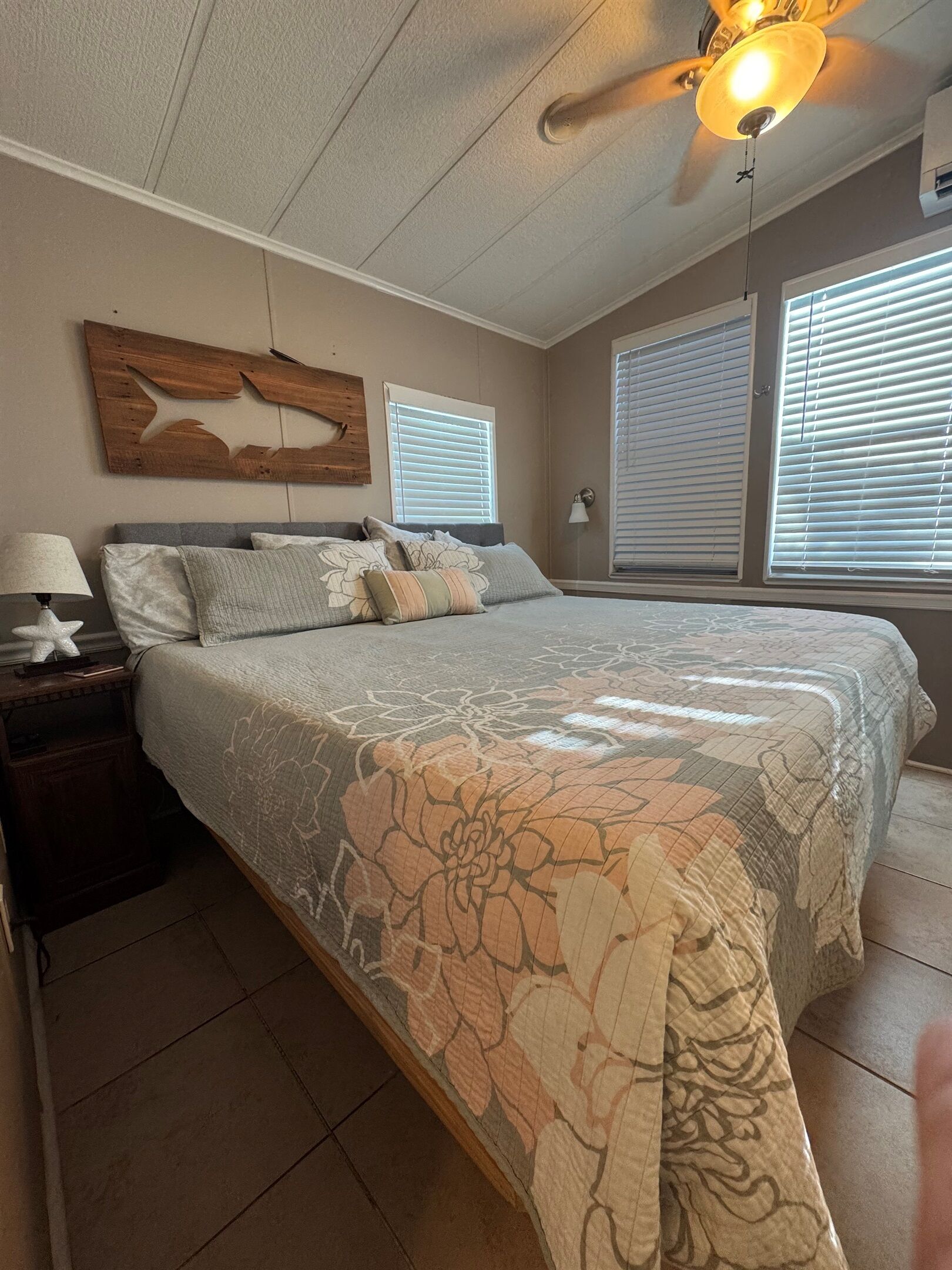 A bedroom with a bed, nightstand, lamp, ceiling fan, and large windows with blinds.